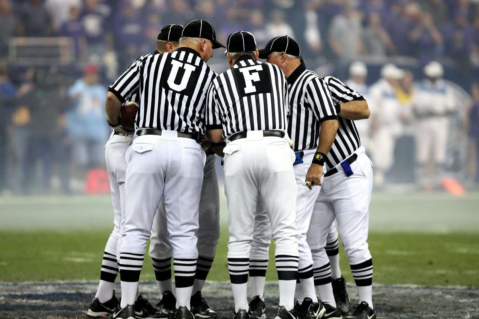 A group of five referees in black and white striped uniforms huddles on a football field, seemingly discussing Section 8 rules for guests as they prepare to enforce fair play and maintain order during the intense match.