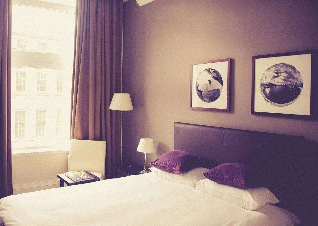 A well-lit bedroom features a double bed with white bedding and purple pillows, two framed artworks above the bed, a floor lamp, and a beige chair next to a window with drawn curtains. This cozy space exemplifies the charm of affordable housing initiatives like those provided by HUD housing programs.