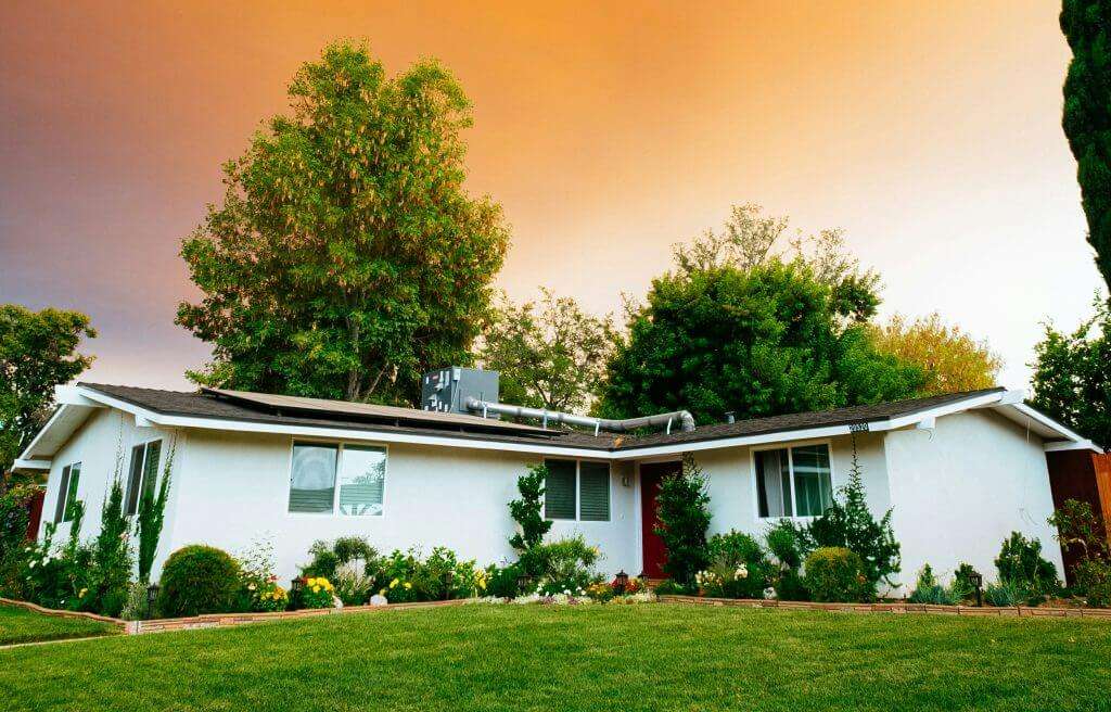 A single-story white house with a dark roof is surrounded by greenery and flowers in the front yard, set against an orange sky at sunset, offering picturesque affordable housing.