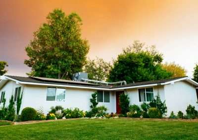 A single-story white house with a dark roof is surrounded by greenery and flowers in the front yard, set against an orange sky at sunset, offering picturesque affordable housing.