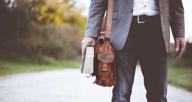 A person standing on a gravel path, holding a book and a brown leather bag, dressed in a gray blazer, white shirt, and dark pants, appears ready to delve into Section 8 facts about affordable housing initiatives.