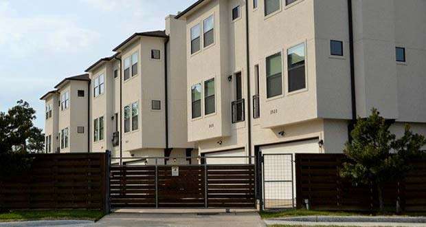 A row of modern three-story townhouses with white exteriors and multiple windows, situated behind a tall wooden fence and metal gate, providing an excellent example of affordable housing.