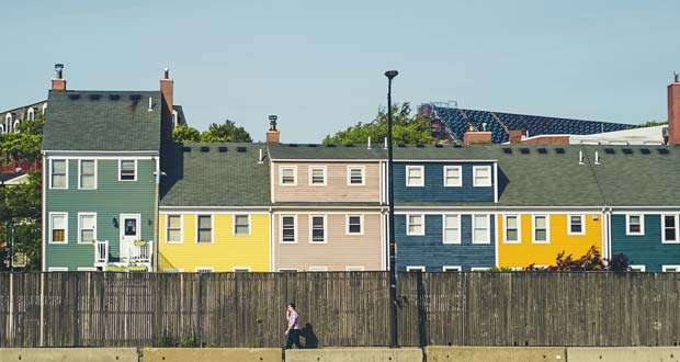 A row of colorful houses behind a wooden fence with a person walking on the sidewalk in front, exemplifying vibrant affordable housing options.