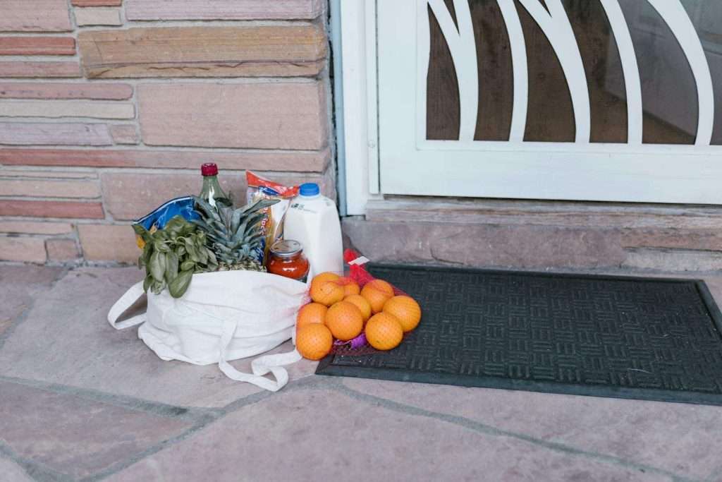 Groceries in reusable bags on a doorstep, including vegetables, fruits, and bottles purchased with food stamps, next to a welcome mat outside a home.