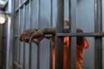 A convicted felon in an orange uniform leans against the bars of a prison cell, with only their arms and part of their upper body visible.