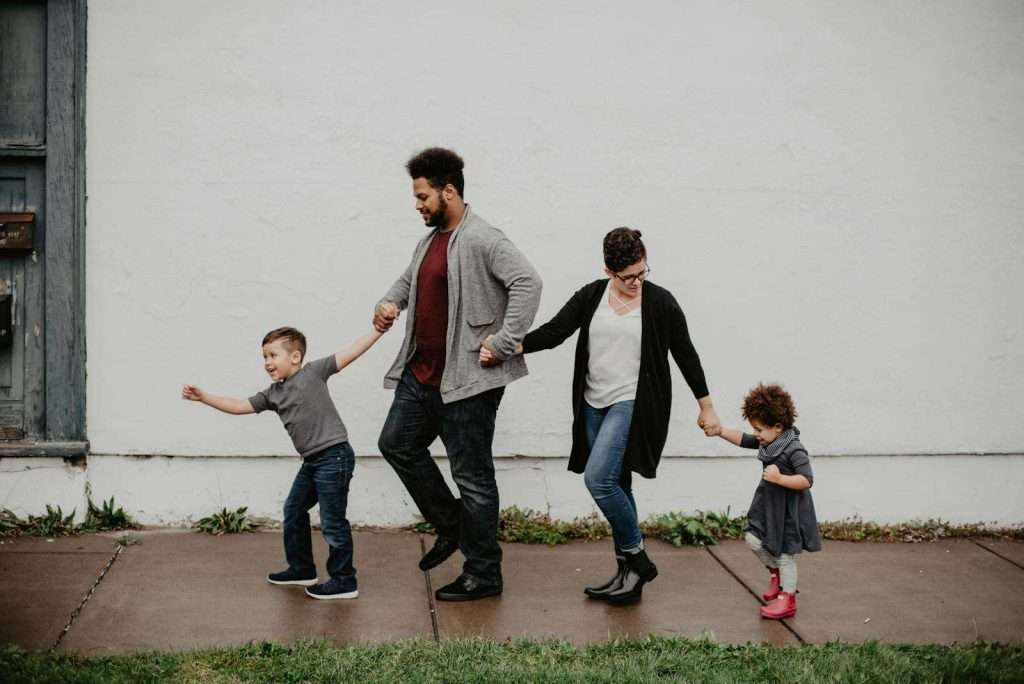 A family of four walks hand-in-hand along a sidewalk; the parents in the middle, with the father holding the son's hand on the left and the mother holding the daughter's hand on the right. Their chatter echoes through Section 8, filled with cozy bedrooms and a close-knit community.