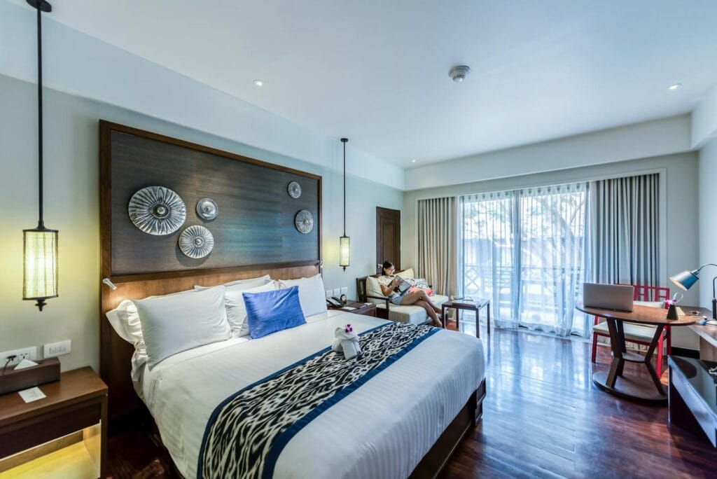 A modern hotel room with a large bed, wooden floor, sofa with a person relaxing, desk, and bright balcony—ideal for guests curious about Section 8 voucher bedroom eligibility or upgrading Section 8 voucher bedroom size.