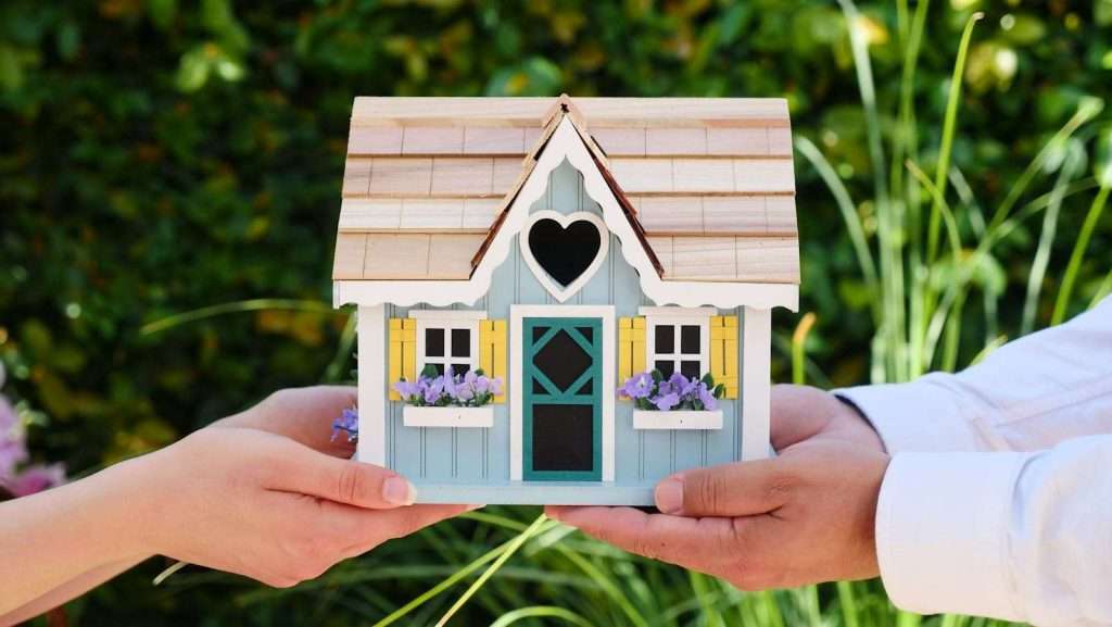Two hands gently cradle a small decorative house adorned with a blue door, heart-shaped window, and flower boxes, evoking the warmth and hope of finding Section 8 housing amidst a lush green leafy background.