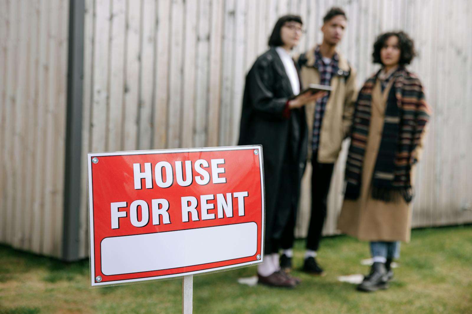 A "House For Rent" sign is in focus in front of three people standing near a wooden fence, one of whom appears to be discussing details with a Section 8 landlord.