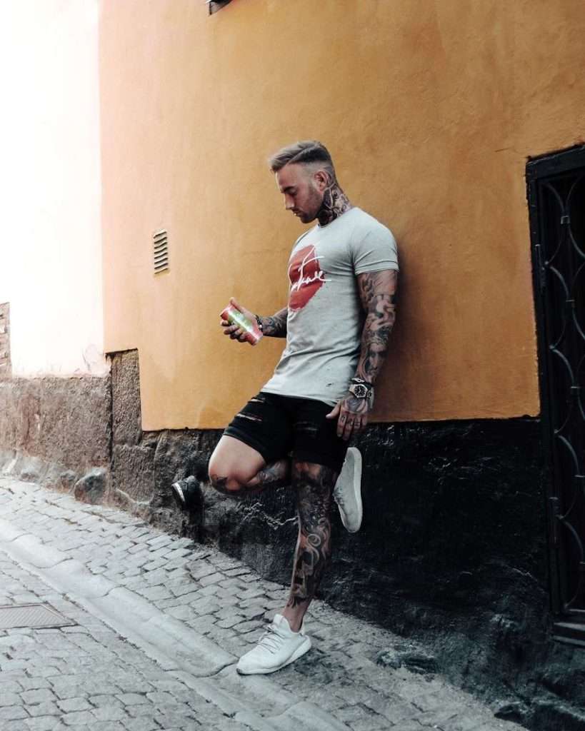 A tattooed man in a grey t-shirt and black shorts leans against an orange wall, sipping from a drink can, quietly reflecting on the section 8 stigma that lingers around him, on a cobblestone street.