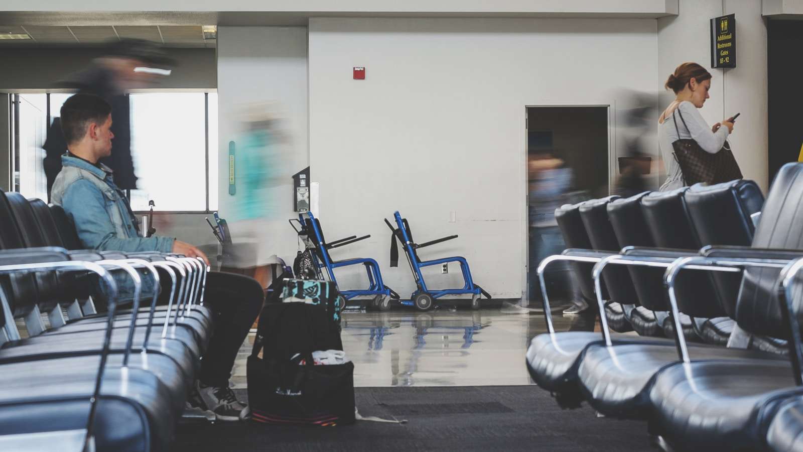The airport waiting area has rows of empty seats, akin to a Section 8 housing waiting list. A few people pass by, one person sits with luggage, and another checks a phone. Two wheelchairs are visible in the background, quietly mirroring the patience often required for such lists.