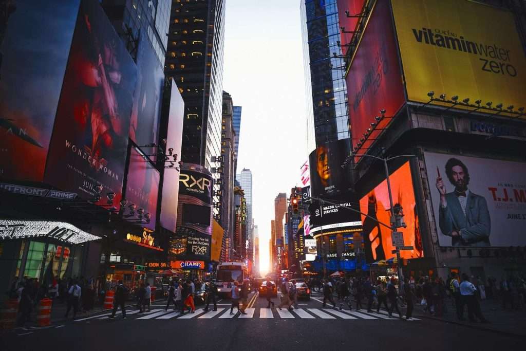 View of a busy New York city intersection, where colorful billboards light up the evening and cars weave through crowds of pedestrians, reflecting the vibrant energy of a city bustling with life under a sunset sky—a scene familiar to those seeking Section 8 housing in New York.